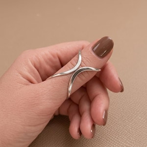May include: A silver ring featuring two curved, smooth bands that meet at the center to form an open, symmetrical shape. The metal has a polished, reflective finish, and the design wraps around the finger with a clean, continuous line.