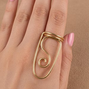 May include: A gold-toned wire ring featuring an elongated, open-frame design that extends vertically along the finger. The metal wire forms a continuous loop with a central, inward-curving spiral detail, creating a bold, linear, and abstract shape.