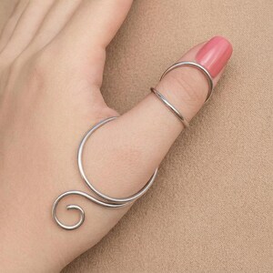May include: A silver-toned metal finger splint featuring a circular band that encircles the thumb, connected to a smaller upper ring that rests on the distal phalanx, finished with a decorative spiral curl at the base of the thumb structure.