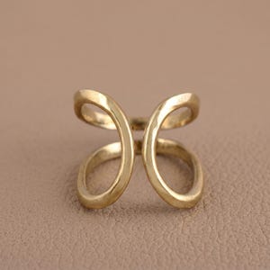 May include: A gold-colored ring with a unique open design. The ring features two curved, looping shapes that meet in the center, creating an abstract, elegant look. The ring is made of a shiny, metallic material.
