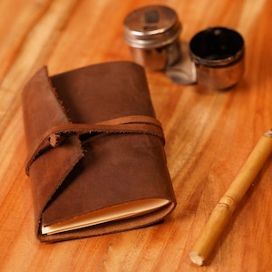 May include: A brown leather journal with a wrap-around tie closure. A bamboo pen with a black tip rests on a wooden surface, along with two small silver containers. The journal is open, revealing blank pages.