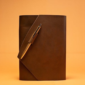 May include: A brown leather journal with a pen holder and a gold-colored pen. The journal has a simple, elegant design with a flap closure. The pen is inserted into a leather loop on the cover. The background is a solid orange color.