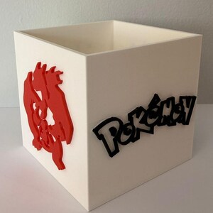 Puede incluir: Un contenedor cuadrado blanco con un gráfico rojo en un lado y la palabra "Pokémon" en negro en otro. El contenedor tiene forma de cubo, con un interior hueco.