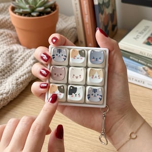 Peut inclure: Un pavé macro pour clavier mécanique blanc et carré comportant neuf touches individuelles, chacune décorée d'un visage de chat de dessin animé unique dans diverses couleurs dont gris, marron, blanc et noir, avec un anneau porte-clés en métal.