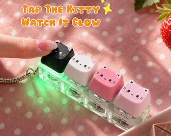 Puede incluir: Una fila de cuatro teclas con forma de gato en negro, blanco y dos tonos de rosa, unidas a un llavero. La tecla negra brilla en verde al presionarla. El texto "Tap The Kitty Watch It Glow" aparece sobre las teclas en letras de color naranja.