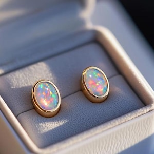 Könnte beinhalten: Ovale Opal-Ohrstecker mit Goldfassungen, präsentiert in einer weißen Schmuckschatulle mit grauem Innenraum. Die Opale zeigen einen farbenfrohen Farbspiel-Effekt.