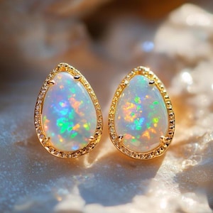 Könnte beinhalten: Ein Paar tropfenförmige Opal-Ohrstecker. Jeder Ohrring hat eine goldene Fassung mit einem Kranz aus kleinen, klaren Steinen. Die Opale zeigen ein Farbspiel mit grünen, blauen und orangen Blitzen.
