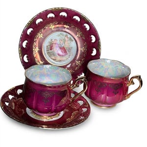 May include: A set of two burgundy teacups and saucers with gold trim and handles. The saucers and a larger plate have a decorative pierced edge. The center of the plate features a painted scene of two people. The teacups have an iridescent interior.