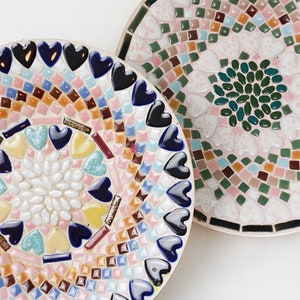 May include: Two round ceramic plates with mosaic designs. One plate has a pink, blue, and brown mosaic with a heart pattern. The other plate has a green, pink, and brown mosaic with a circular pattern.