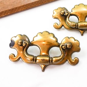 May include: Two antique brass drawer pulls with ornate, symmetrical designs. Each pull features a central handle with decorative flourishes on either side. The pulls are a warm, aged brass color, suggesting vintage charm.