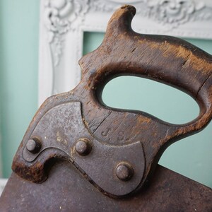 May include: Close-up of an antique hand saw with a wooden handle. The handle is dark brown and has a unique shape. The metal blade is rusty and has a J.S. marking.