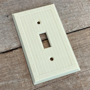 May include: Off-white, rectangular light switch cover with a single toggle opening and two screw holes. The cover has a vertical striped pattern and a decorative border. The switch plate is made of a solid material and is set against a wooden background.