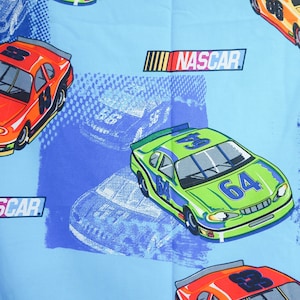 May include: A blue fabric with a repeating pattern of race cars. The cars are green, orange, and blue with black numbers. The text "NASCAR" is printed in black on the fabric.