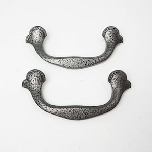 May include: Two silver-toned metal drawer pulls with a decorative, slightly curved design. The pulls have a textured, slightly rough finish.