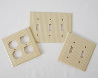 May include: Three ivory-colored plastic electrical outlet covers with a ribbed texture. Two covers have single light switch openings and one cover has two electrical outlet openings.
