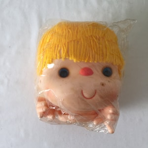 May include: A plastic doll head with yellow yarn hair, blue eyes, and a red nose. The doll is packaged in clear plastic.