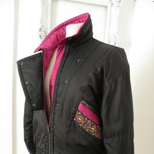 May include: A black quilted jacket with a pink lining and a colorful embroidered patch on the pocket. The jacket has a zipper closure and a ribbed hem.