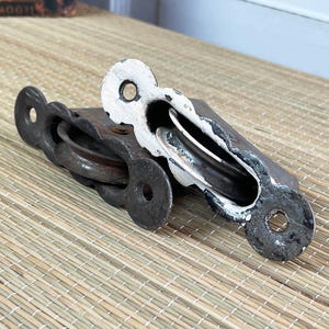May include: Two antique metal pulleys with a distressed, rustic appearance. The pulleys have a dark, weathered finish with white paint chipping off in areas. The design includes a wheel and a hole for mounting. Ideal for vintage decor or restoration projects.