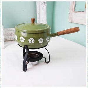 May include: Green enamel fondue pot with a white floral design and a wooden handle, sitting on a black metal stand with a small metal bowl.
