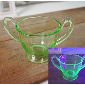 May include: A green glass sugar bowl with two handles. The bowl is made of uranium glass and glows under a black light.