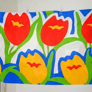 May include: A rectangular fabric with a vibrant floral pattern. Large red and yellow tulip-like flowers with green stems and leaves are set against a white and blue background. The fabric is hung with white clips.