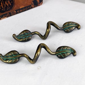 May include: Two antique brass leaf-shaped drawer pulls. Each pull features a curved stem and a leaf with green detailing. The hardware is designed to resemble a vine with leaves. The pulls are on a white surface.