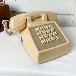 May include: A vintage AT&T 100 push-button telephone in a cream color. The phone features a keypad with white buttons and a beige center panel. The handset is connected with a coiled cord. The phone is a retro home decor item.