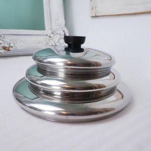 May include: A stack of three stainless steel pot lids with a black handle on top. The lids are circular and have a shiny, reflective surface. The lids are nested together, with the largest lid at the bottom.