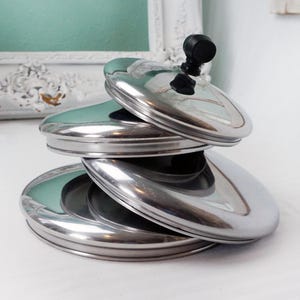 May include: A stack of four shiny, stainless steel pot lids. Each lid has a rounded, oval shape with a reflective surface. The top lid features a small, black, round handle. The lids are stacked on top of each other, creating a modern, minimalist aesthetic.