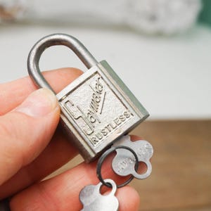 May include: A vintage silver-colored padlock with a curved handle and the word "Rustless" printed on the front. The padlock has a key attached to a small ring. The brand name "Slaymaker" is also visible on the lock.