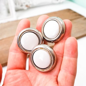 May include: Three white ceramic knobs with silver metal trim. The knobs are round and have a smooth, glossy finish.