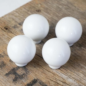 May include: Four white, spherical cabinet knobs with a glossy finish are arranged on a weathered, wooden surface. Each knob has a small, rounded base. The knobs are evenly spaced, creating a symmetrical pattern against the rustic background.