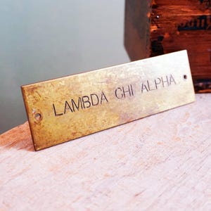 May include: A rectangular brass plaque with the engraved text "LAMBDA CHI ALPHA". The plaque has two small holes and a slightly aged appearance. The background includes a wooden box and a light-colored surface.