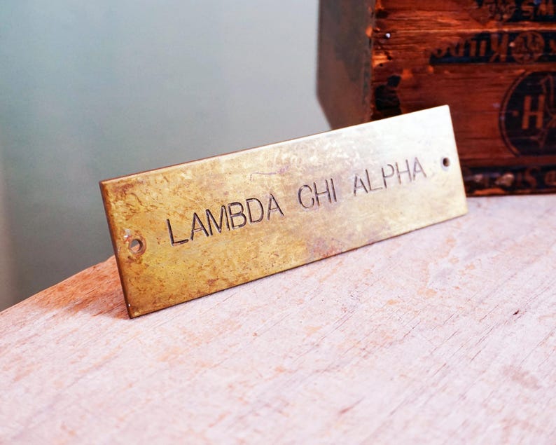Pode incluir: Placa retangular de lat&atilde;o com o texto gravado "LAMBDA CHI ALPHA". A placa tem dois pequenos orif&iacute;cios e uma apar&ecirc;ncia ligeiramente envelhecida. O fundo inclui uma caixa de madeira e uma superf&iacute;cie clara.