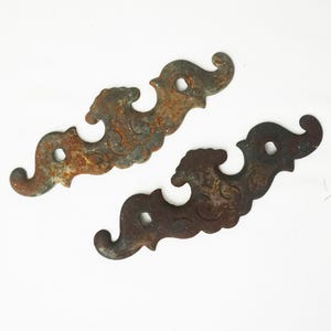May include: Two antique, decorative metal brackets with a distressed, rusty finish. Each bracket features an ornate, swirling design with a central hole for mounting. The brackets are a dark brown color with areas of orange rust.
