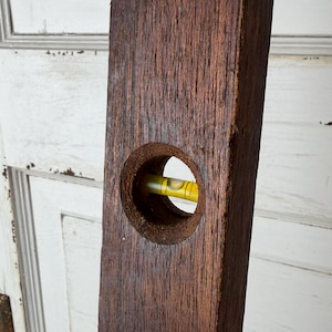 May include: A vintage spirit level tool constructed from dark brown, textured wood. The tool features a circular cutout in the center, which houses a small, horizontal glass vial filled with yellow liquid and a single air bubble for leveling.