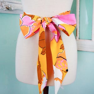 May include: A colorful floral bow belt in shades of orange, pink, brown, and white. The belt is tied in a large bow, with long, flowing tails. The fabric features a bold, retro-inspired floral pattern. The belt is displayed on a mannequin.