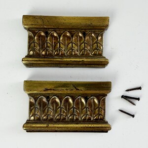 May include: Two rectangular brass-toned metal furniture mounts featuring a repeating embossed leaf pattern along the center. Each piece has a flat top and bottom edge. Four small, dark metal nails are included for installation purposes.