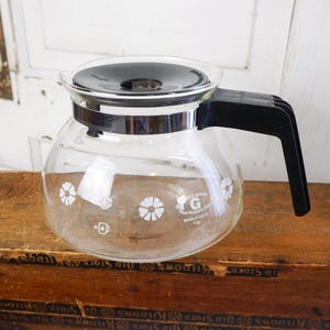 May include: A vintage clear glass coffee pot with a black handle and lid. The pot has a silver band around the middle and white floral designs. The text "G Brand" and "Made in USA" are visible on the glass.