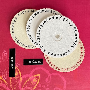 May include: A collection of cream and white letter and number wheels with black and red lettering. The wheels are arranged on a pink background with the word "etsy" in black. The word "ETSY" is also displayed vertically.