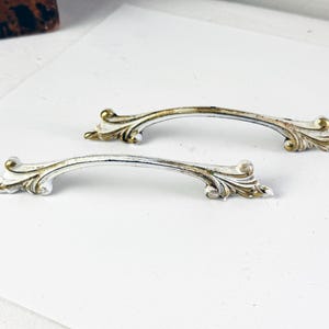 May include: Two vintage cabinet pulls with a white and gold finish. The pulls have a curved design with ornate detailing on the ends. The hardware is likely made of metal and is designed for furniture.