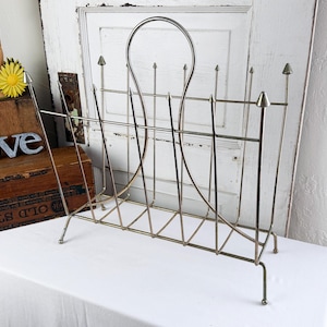 May include: A vintage metal magazine rack with a silver finish. The rack features a curved handle and vertical bars with decorative arrow-shaped finials. The design is open, with a geometric pattern at the base. The rack is on a white surface.