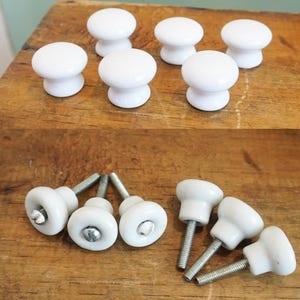 May include: A collection of white ceramic cabinet knobs with a rounded top and a cylindrical base. Some knobs are shown with their metal screws attached. The knobs are displayed on a wooden surface.