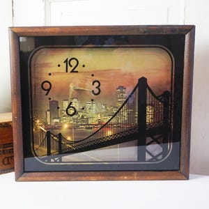 May include: A square wall clock with a dark brown wooden frame. The clock face features a nighttime cityscape with a bridge and city lights. The clock hands point to 10:10.