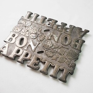 May include: A silver-toned metal trivet with the words "Bon Appetit" and "Lilly" in a decorative font. The trivet is decorated with a floral design of strawberries.