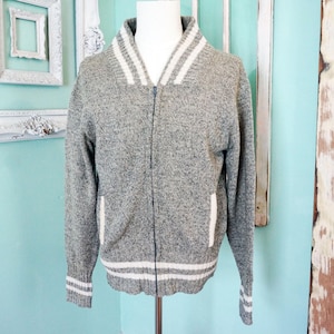 May include: A gray zip-up sweater with white stripes on the collar, cuffs, and hem. The sweater has a classic design with a full zipper and two front pockets. The fabric appears to be a knit material, giving it a cozy look.