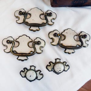 May include: A collection of antique drawer pulls and knobs. The pulls have a white, cloud-like design with ornate, dark metal handles. The knobs are smaller, with a similar design. All pieces are set against a white background.