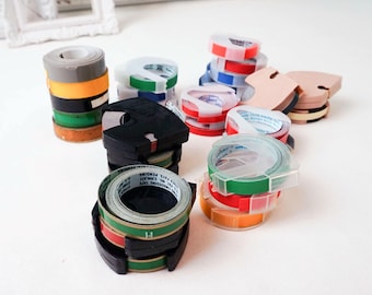 May include: A collection of colorful label tapes in various colors, including green, red, orange, and blue. The tapes are stacked and organized in clear plastic dispensers and black holders. Some tapes have printed text.