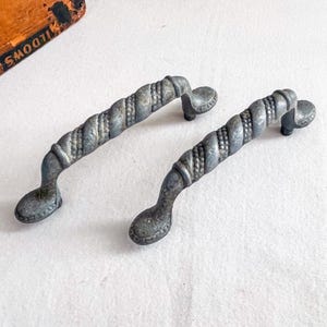May include: Two antique, silver-toned cabinet pulls with a twisted rope design. The pulls have a decorative, oval-shaped base and are likely made of metal. These vintage handles would be suitable for a dresser or cabinet.