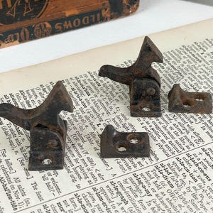 May include: Four antique, rusty metal hinges and brackets, likely from a cabinet or chest, are arranged on a vintage book page. The hardware has a weathered appearance, suggesting age and use.